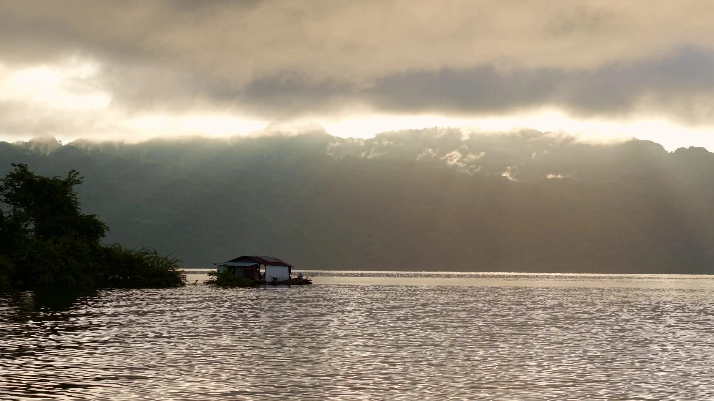 A wooden floating house at dusk on Khao Laem Lake, ringed by forested mountains.