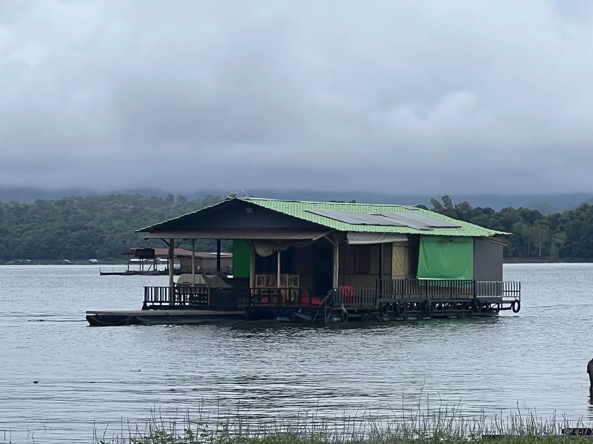 A green-roofed wooden raft house moored on the still water of Khao Laem Lake, forested hills behind.