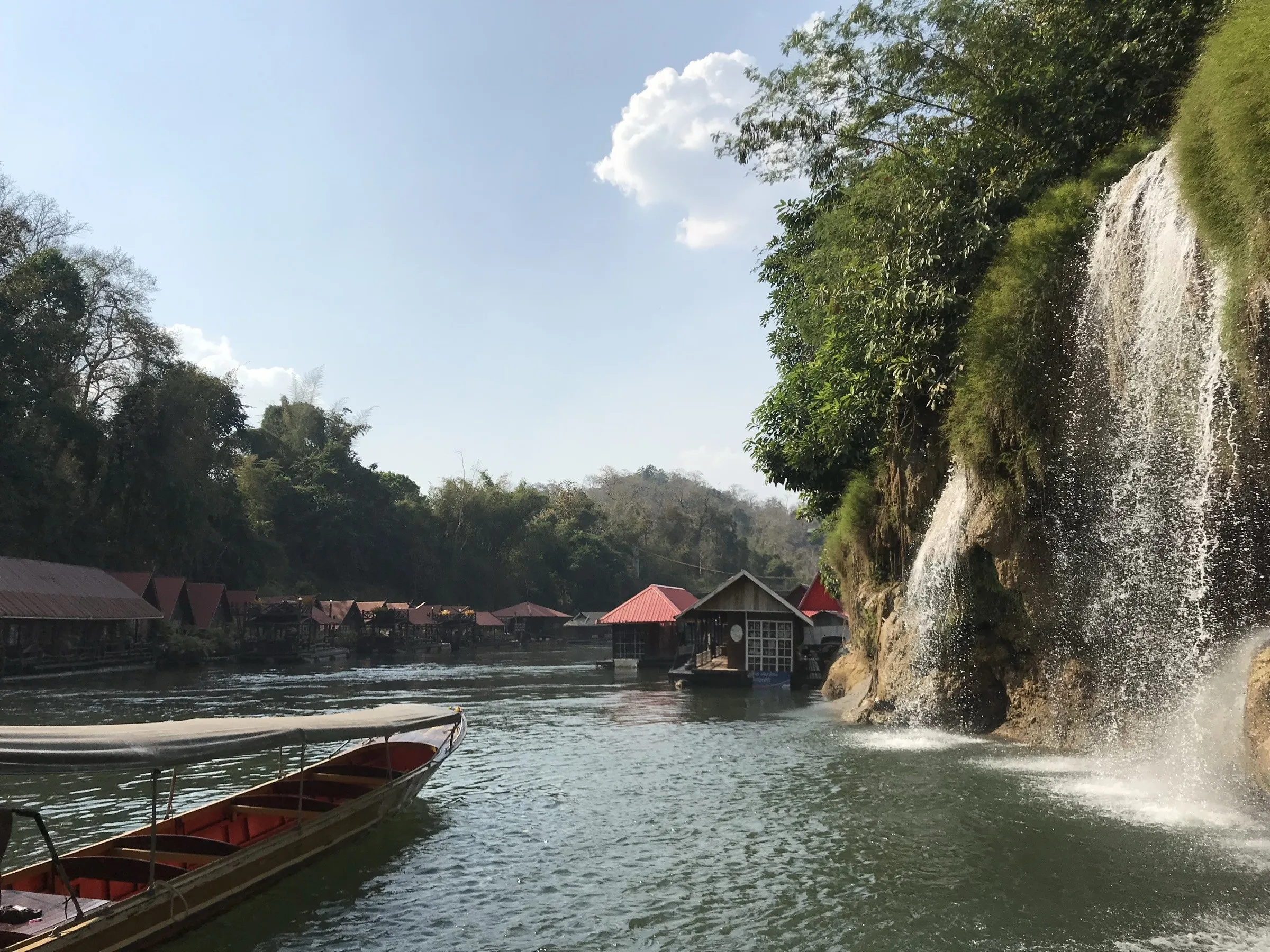 Wooden raft houses and a waterfall on the River Kwai at Sai Yok, a longtail boat moored in the foreground.